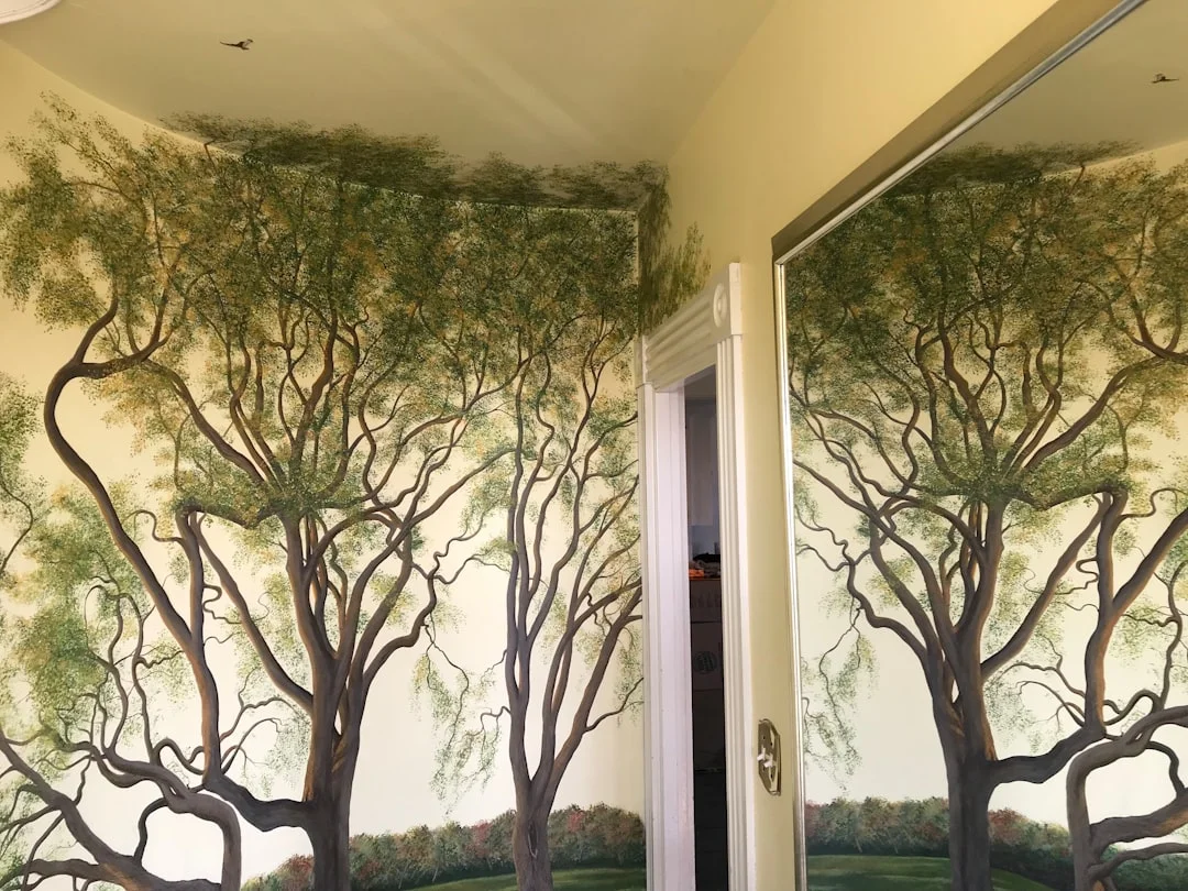 A sun-filled principal bedroom with floor-to-ceiling hand-painted chinoiserie wallpaper panels in soft jade and cream, depicting blossoming trees and perched birds, with a linen upholstered bed in the foreground